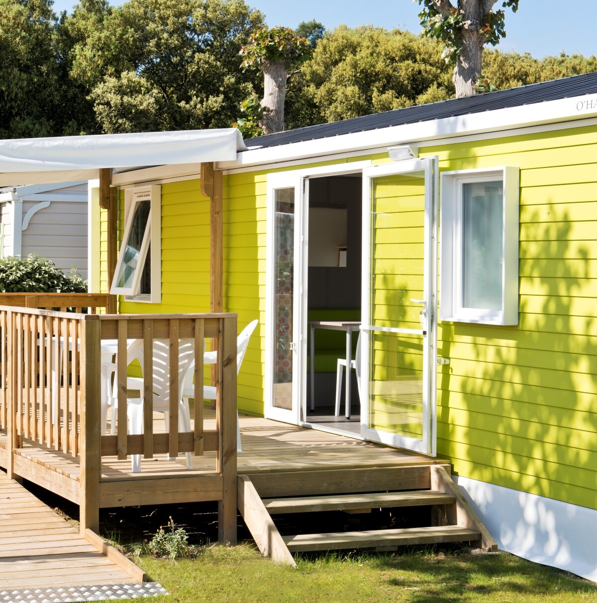 A bright yellow mobile home features a wooden terrace with outdoor seating. The entrance is framed by steps leading up to the door, which opens to a light-filled interior. Nearby greenery enhances the natural setting.