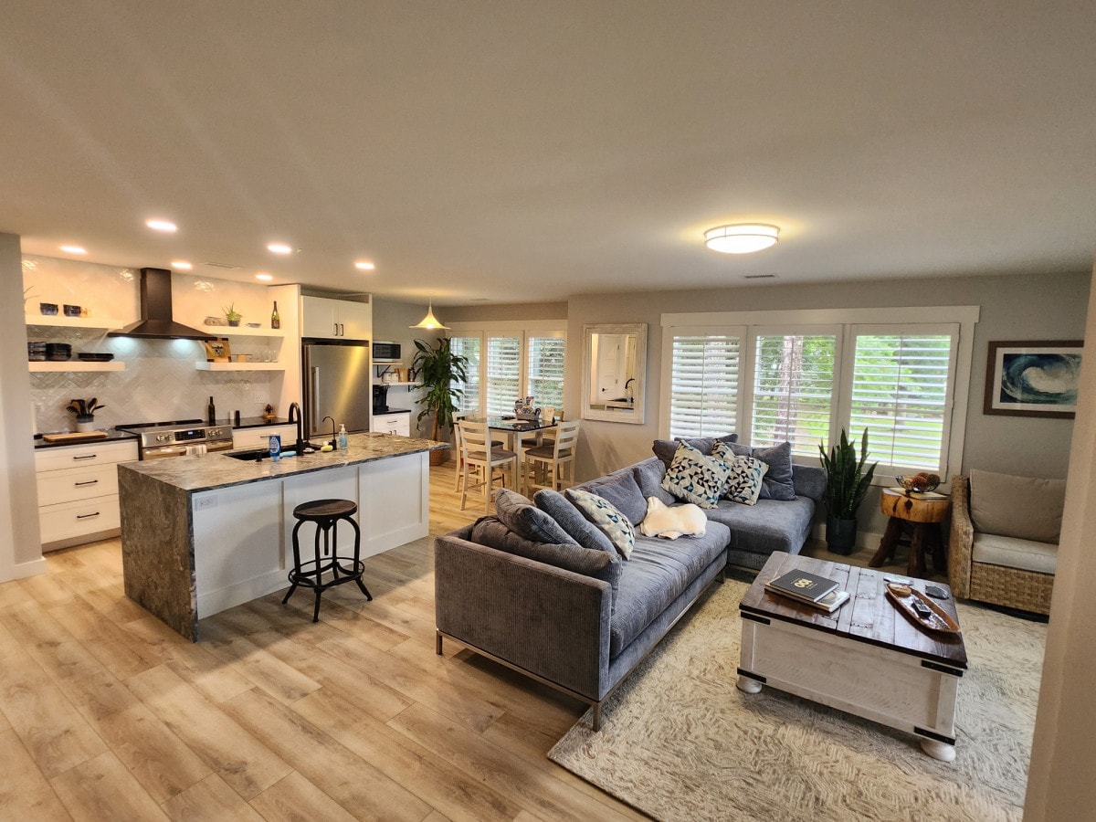 An open living space features a spacious grey sofa with assorted cushions, a cozy coffee table, and a modern kitchen equipped with stainless steel appliances. Sunlight filters through plantation shutters, illuminating the inviting dining area with a wooden table and chairs. Lush green plants are placed throughout.