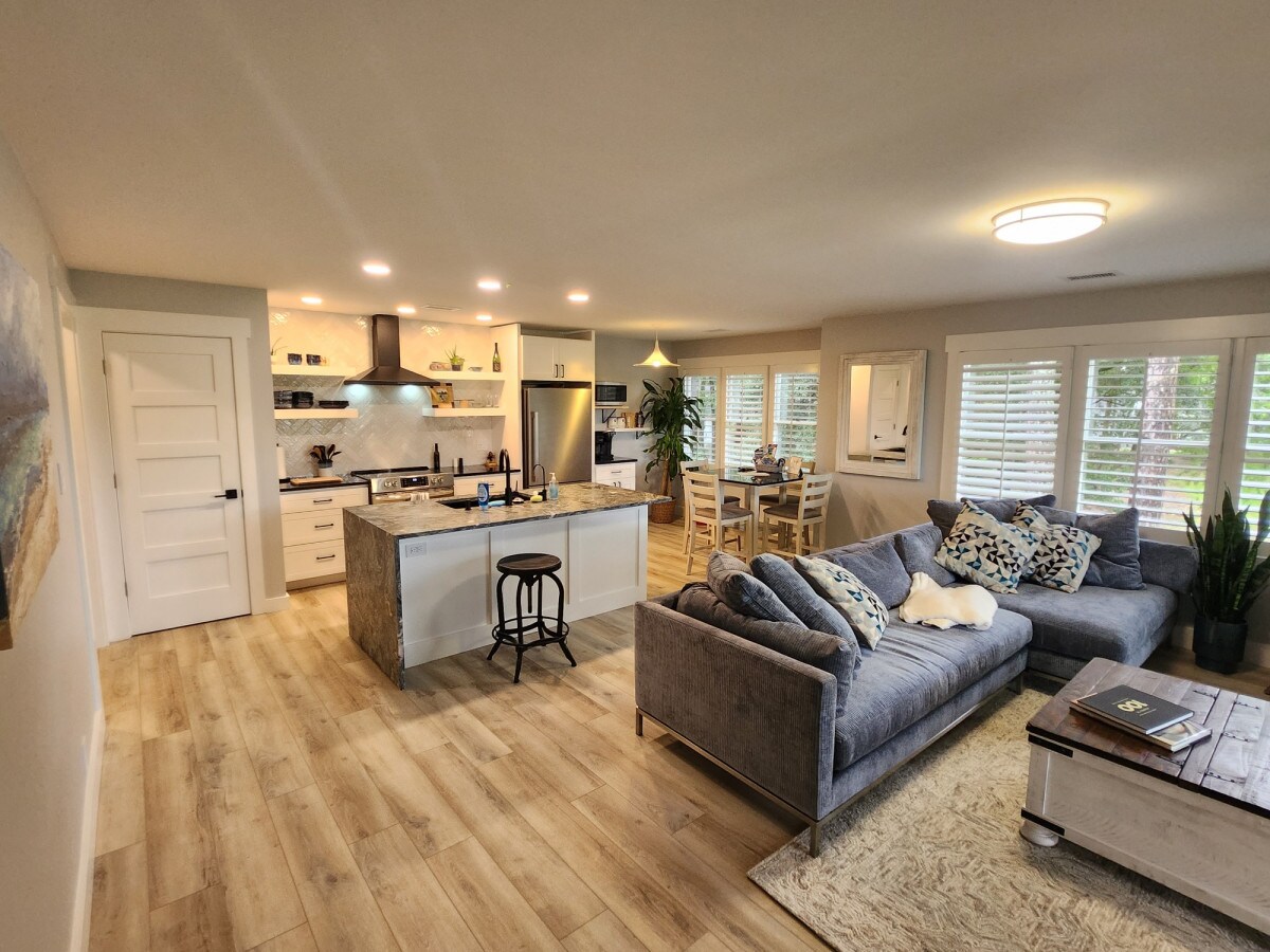 An open-concept living space features a modern kitchen with bright cabinetry and an island, inviting casual dining. A plush sectional sofa is arranged comfortably in the living area, accompanied by a decorative coffee table. Natural light floods in through window treatments, enhancing the cozy ambiance.
