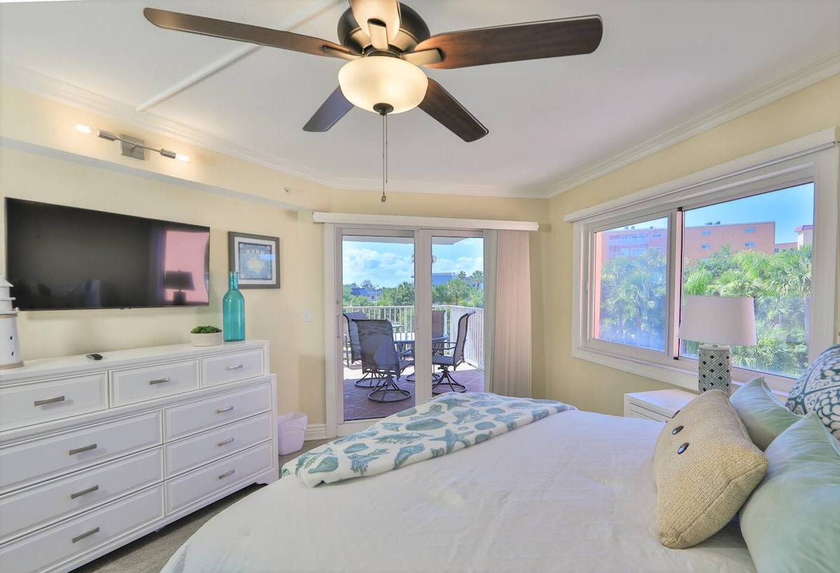 A bright and airy bedroom features a spacious bed adorned with decorative pillows. A flat-screen TV is mounted above a dresser. Large windows provide natural light and access to a private balcony with seating, overlooking lush palm trees.