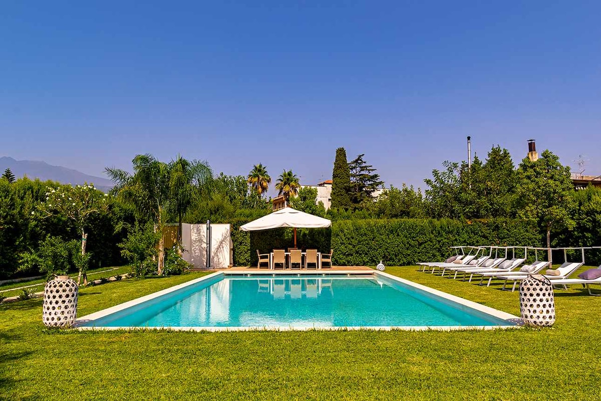 A spacious swimming pool sits surrounded by a well-maintained lawn, featuring sun loungers neatly arranged on one side. A shaded seating area with an umbrella provides a comfortable spot for relaxation, while lush greenery frames the area, enhancing the tranquil ambiance.