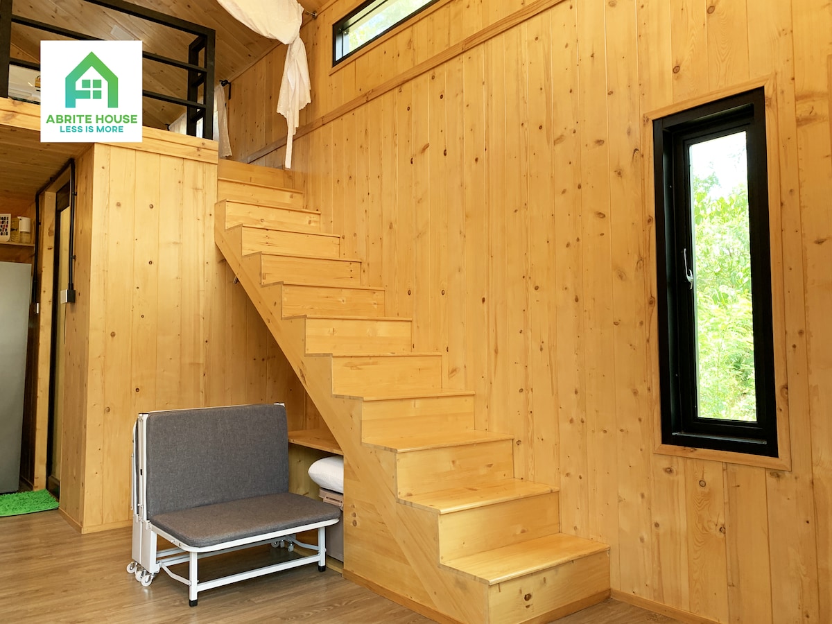 The interior features warm wooden walls and a staircase leading to the mezzanine. A foldable seating area is positioned against the wall, providing additional space. Natural light enters through a nearby window, enhancing the inviting atmosphere of the living area.