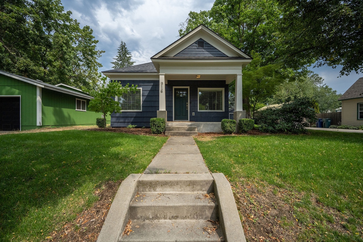 A charming exterior view of the house showcases a well-maintained front yard with a concrete pathway leading to the entrance. The home's facade features blue siding, white columns, and large windows, complemented by landscaped greenery and a spacious lawn.