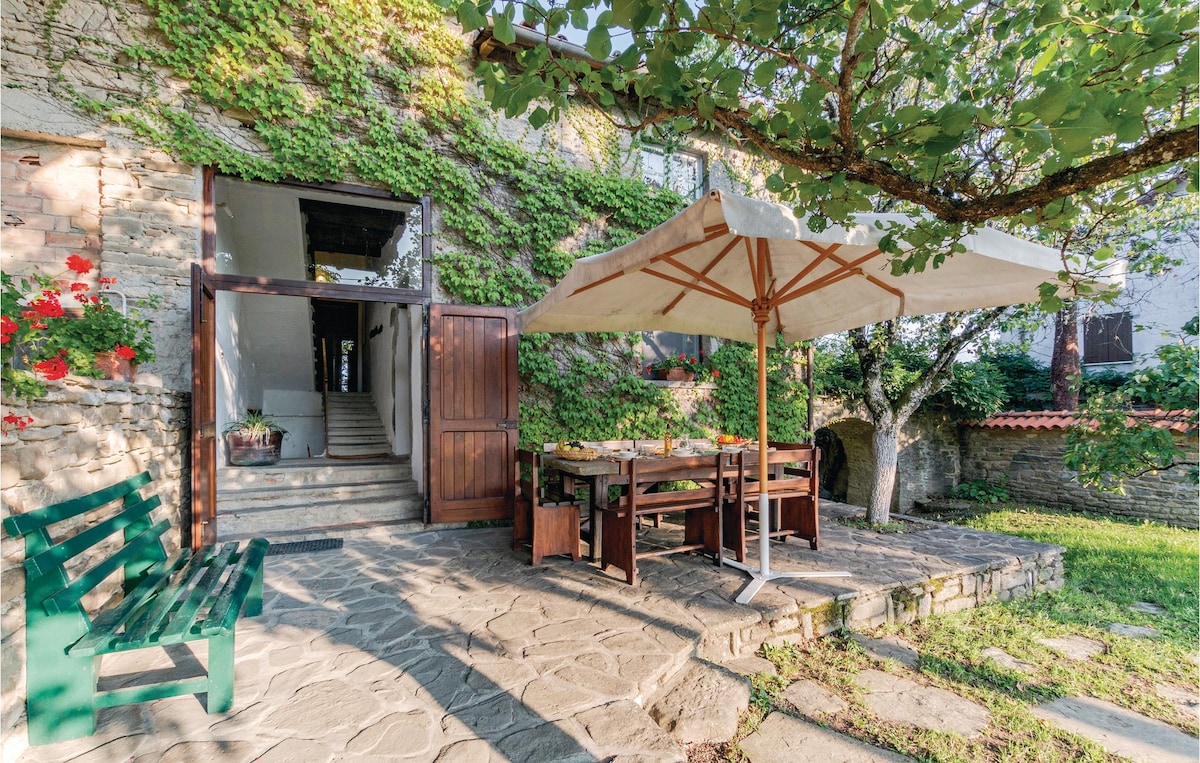 The outdoor dining area is featured under a large umbrella, surrounded by lush greenery. A wooden table set with chairs is positioned on a stone patio, with steps leading up to the entrance of the house. A climbing plant enhances the natural scenery.