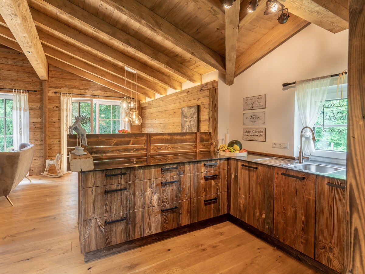 The kitchen area features wooden cabinetry complemented by a dark countertop. Large windows provide natural light, highlighting wooden beams on the ceiling. A fruit arrangement is visible, along with modern kitchen appliances. The space is designed with an open layout, facilitating a seamless transition to the adjacent living area.