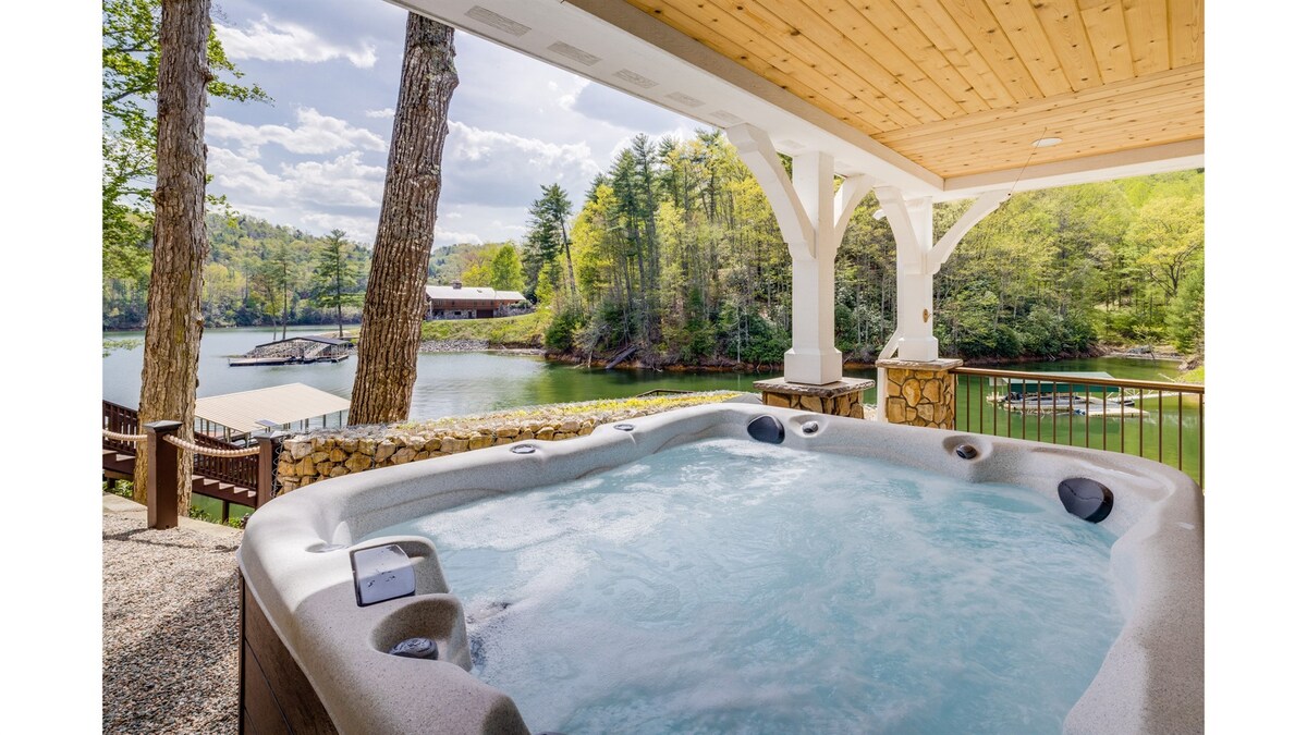 A hot tub is situated on a covered porch, overlooking a tranquil lake surrounded by trees and scenic hills. The inviting water reflects the natural beauty of the cove, creating a peaceful atmosphere for relaxation.