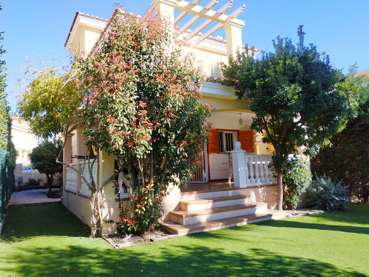 A charming chalet is surrounded by well-maintained greenery and vibrant flowering plants. A small porch leads to the entrance, framed by decorative columns. The spacious lawn is complemented by neatly trimmed shrubs, creating a tranquil outdoor space. Sunlight brightens the scene under a clear blue sky.