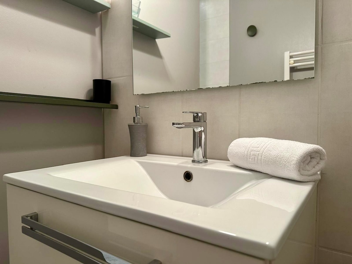 The bathroom features a modern vanity unit with a spacious white sink. A clean rolled towel is placed beside a soap dispenser, and a mirror extends above the sink. Minimal shelving is visible, completing the functional design.