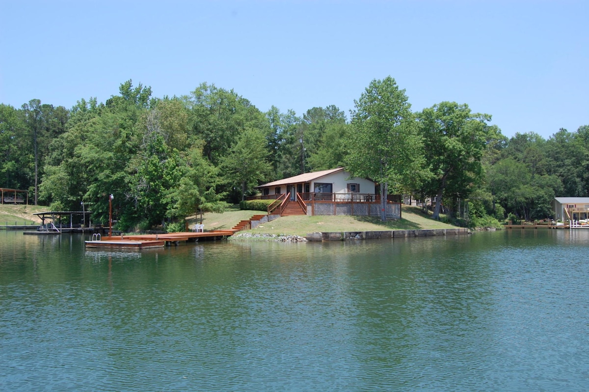 A lakefront cabin is set against a backdrop of lush greenery, with a gently sloping yard leading to the water. A wooden dock is positioned alongside the cabin, providing access to the lake. Clear blue skies reflect off the calm water.