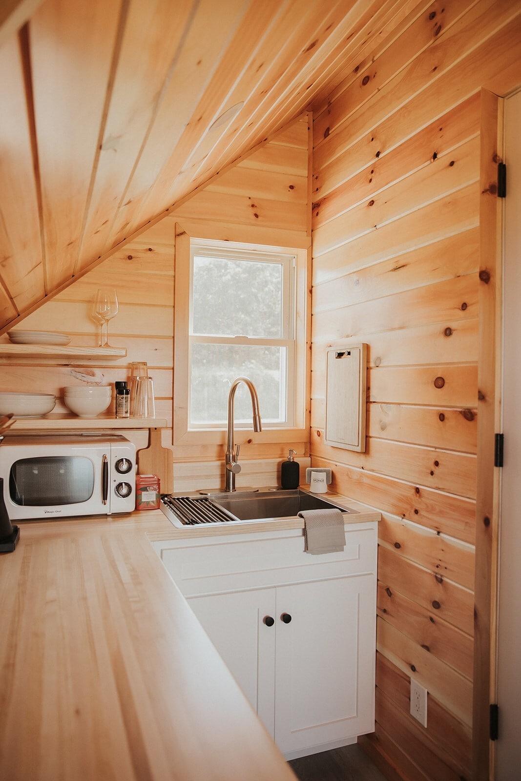 The kitchenette features warm wooden walls and a sloped ceiling, highlighting the natural aesthetic. A stainless steel sink sits alongside a mini fridge and microwave. Minimalist white cabinetry and a small window allow natural light to brighten the space, creating a functional and inviting area.