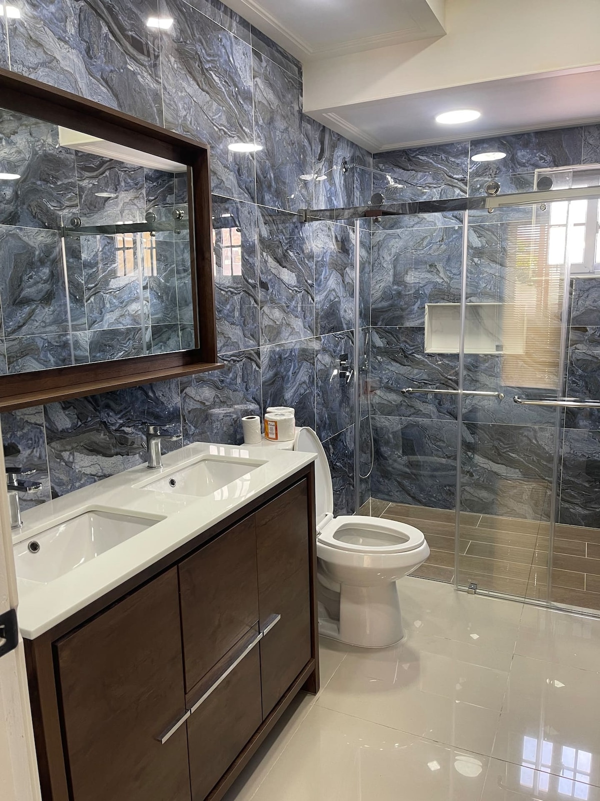A modern bathroom features elegant gray marble walls and a contrasting wooden vanity with dual sinks. A glass-enclosed shower is visible, showcasing sleek fixtures and minimalistic design. Bright lighting enhances the spacious feel, while a large mirror completes the contemporary aesthetic.