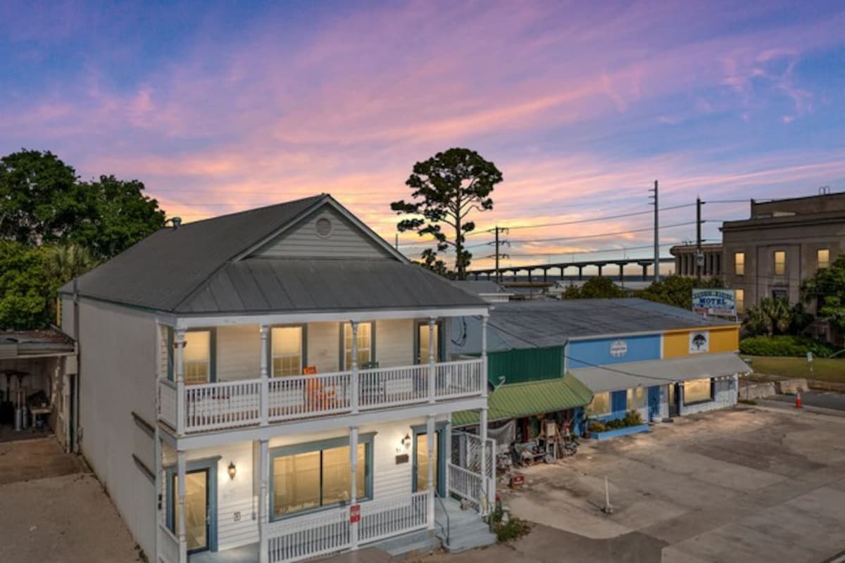 An exterior view of the two-story building showcases a charming design against a vibrant sunset sky. Balconies line the front of the building, with multiple windows allowing natural light into the apartments. Nearby, shops and restaurants are visible along the street.