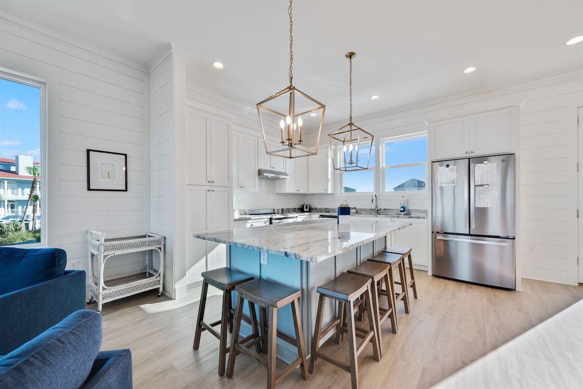A modern kitchen showcases a large island with seating for five, surrounded by white cabinetry and granite countertops. Natural light fills the space through multiple windows, illuminating stainless steel appliances, including a refrigerator and oven. A comfortable seating area is visible nearby.