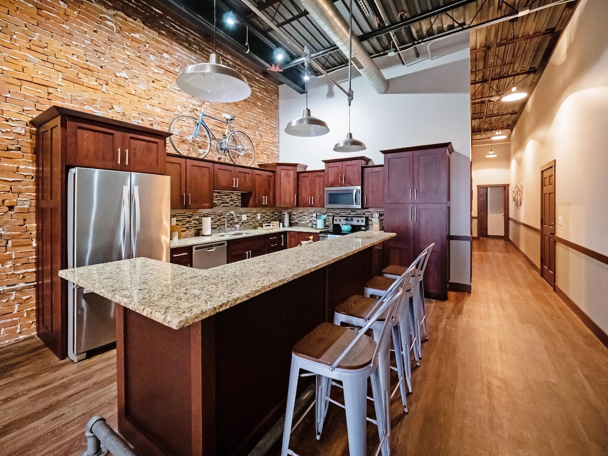 A modern kitchen features dark wood cabinetry and granite countertops, creating a sleek workspace. Stools provide seating along a spacious island. Exposed brick walls contribute to the industrial charm, while overhead pendant lights illuminate the area. A bicycle decorates the wall above, adding a whimsical touch.