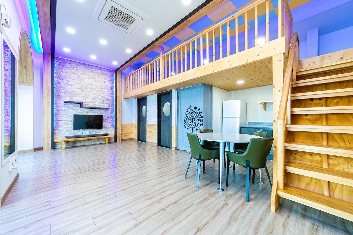 A spacious living area features wooden accents and a lofted sleeping space. A large flat-screen TV is mounted on the wall, while a dining table is set with comfortable chairs. Natural light fills the room through large windows, creating an open and inviting environment.