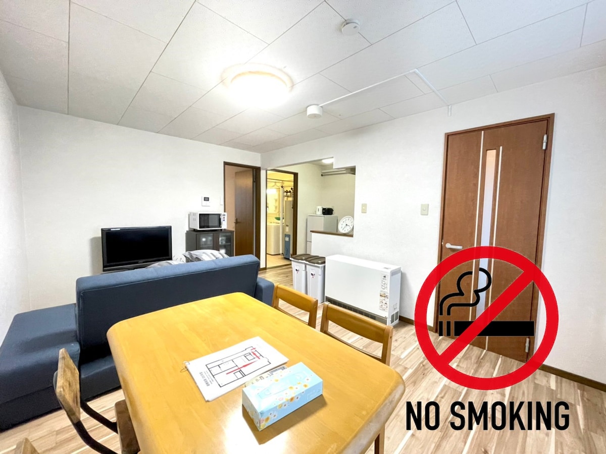 A cozy living area features a dark blue sofa and a wooden dining table with chairs. A television is positioned on a cabinet, while a kitchen can be seen in the background. A no smoking sign is displayed prominently.