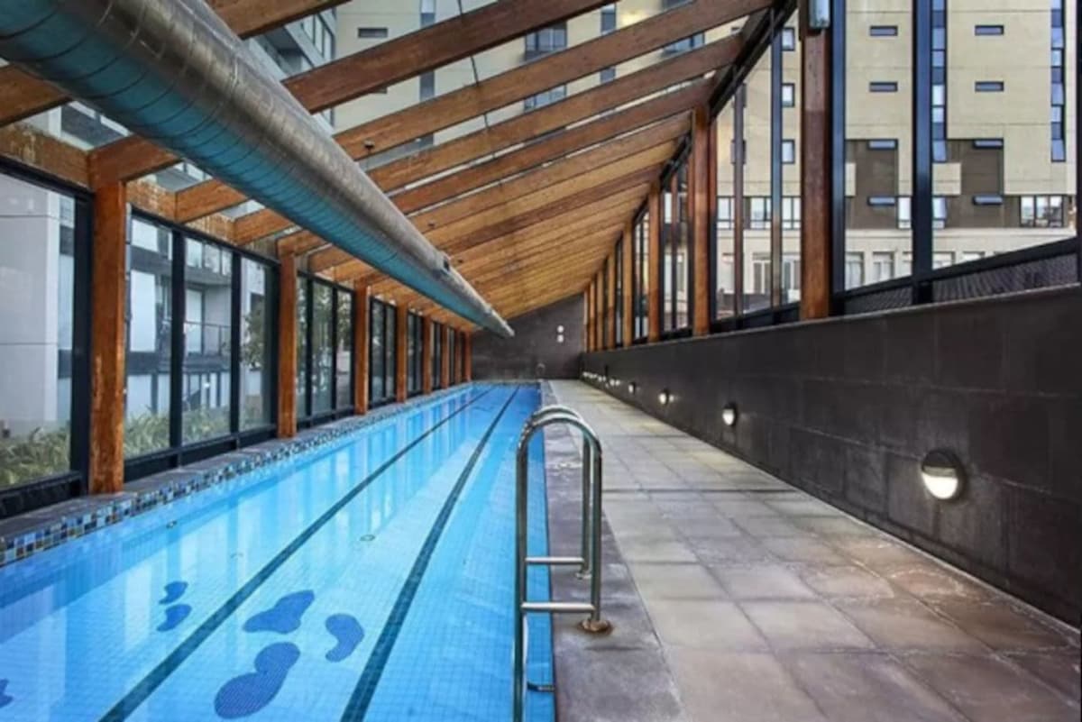 An indoor swimming pool is framed by sleek glass walls and wooden beams. The pool features a clear blue surface with gentle patterns, flanked by a tiled pathway and modern lighting along the walls, creating a relaxed atmosphere.