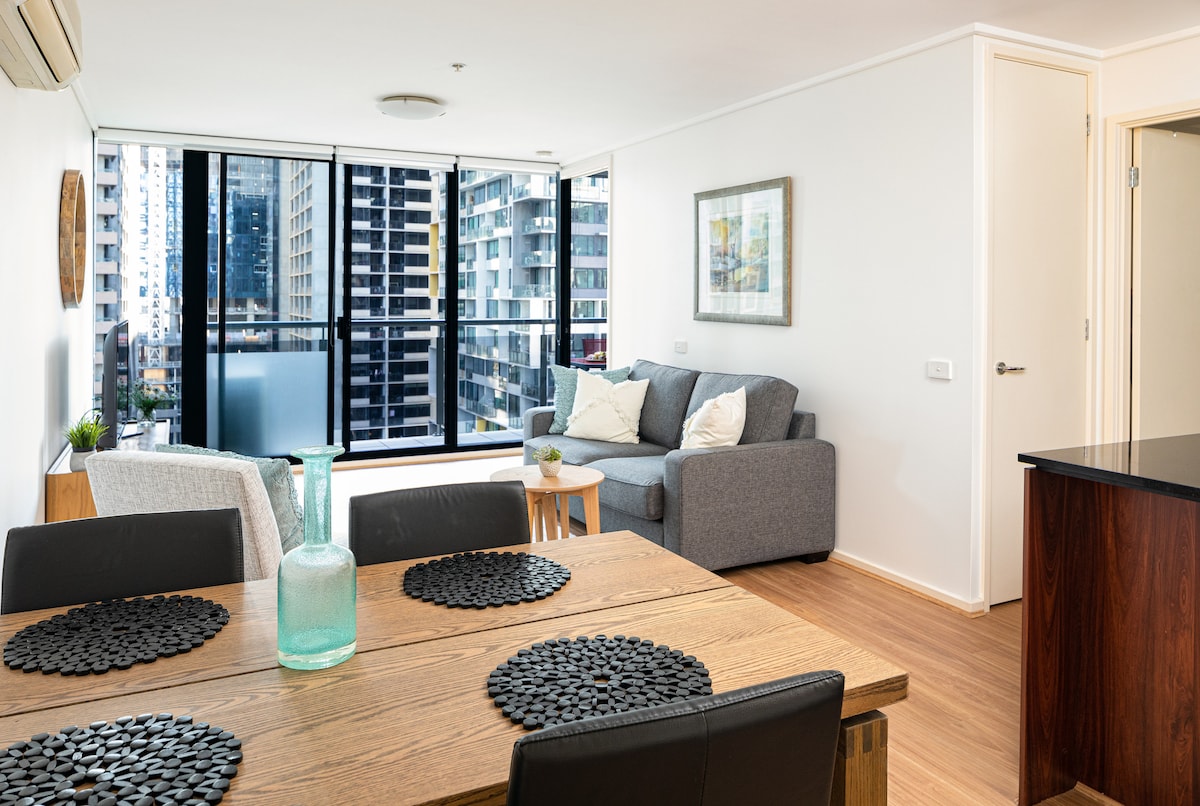 An open-plan living space is shown, featuring stylish floorboards and large floor-to-ceiling windows that offer city views. A dining table with placemats is accompanied by black chairs. The living area contains a gray sofa, a small side table, and a decorative piece on the wall.