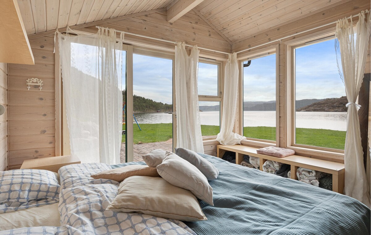 A bright bedroom is seen featuring large windows that offer a view of the fjord and nature outside. The bed is adorned with layered pillows and a soft blanket, while light curtains frame the windows, allowing natural light to fill the room.