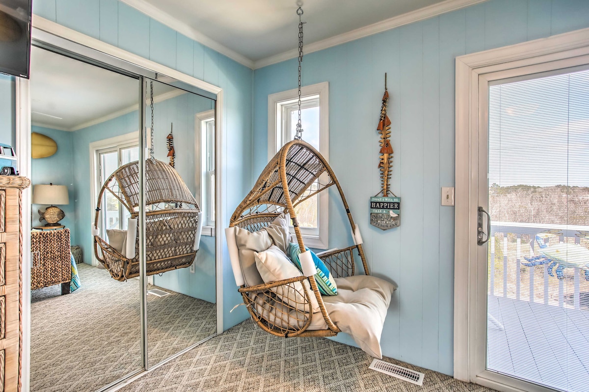 A comfortable hanging chair is featured in a bright blue room, complemented by light-colored walls and ample natural light. A large mirror reflects the chair and nearby windows, enhancing the open feel. Decorative elements include a nautical-themed sign and soft cushions.