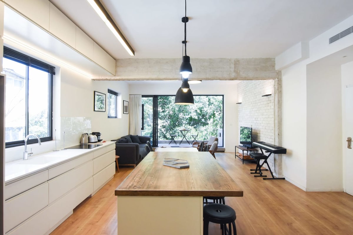 An open-plan kitchen blends with the living area, featuring a wooden island with seating. Large glass doors lead to a balcony, offering views of greenery. A sofa and a small table are situated nearby, alongside a television and a workspace with a keyboard.