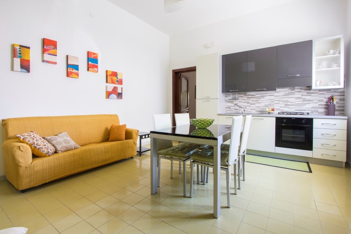 A bright and airy kitchen area features a dining table with six chairs, complemented by a cozy sofa. Colorful artwork adorns the wall, adding a touch of character. Modern appliances are visible in the functional kitchen space, designed for convenience and comfort.
