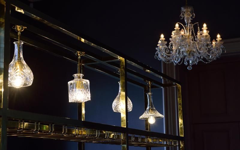 A display of elegant glass light fixtures hangs from a stylish metallic rack, showcasing various shapes and sizes. A glittering chandelier with ornate details is suspended above, illuminating the area with soft light against a dark background.