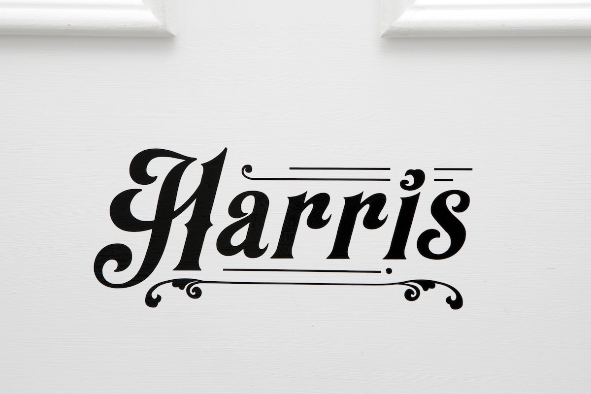 A decorative sign featuring the name 'Harris' is displayed on a white door. The lettering is styled in an elegant, vintage font, providing a classic touch to the entrance.
