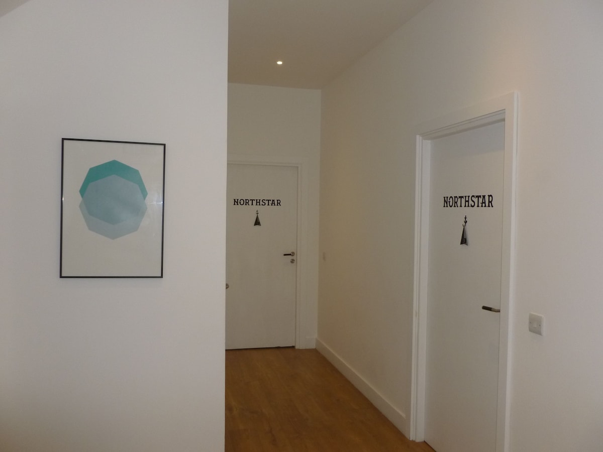 A simple hallway is presented, featuring two white doors marked 'NORTHSTAR.' A framed geometric artwork in subtle blue tones is mounted on the wall. The warm wooden flooring extends throughout the space, contributing to a clean and modern aesthetic.