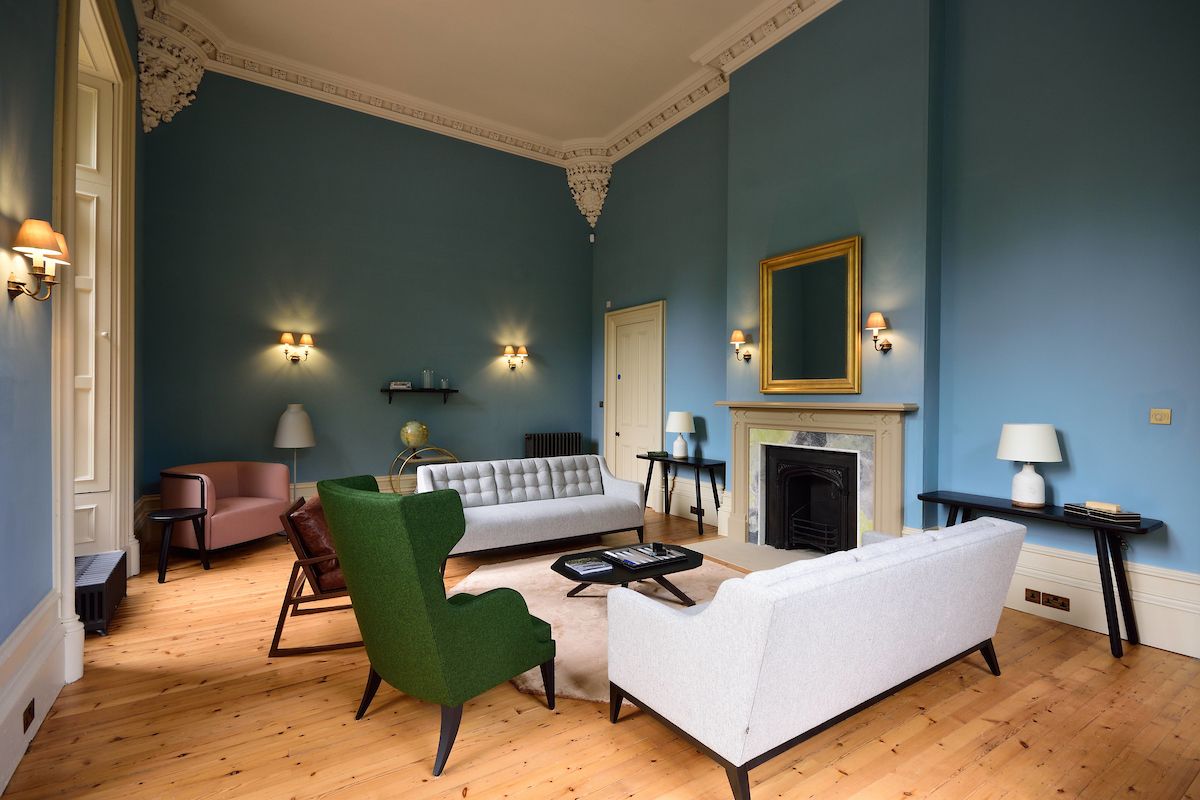 A spacious living area features high ceilings and elegant moldings. A mix of modern and classic furnishings includes a green accent chair, gray sofas, and side tables. Soft lighting is provided by wall sconces and table lamps, creating a calm ambiance.
