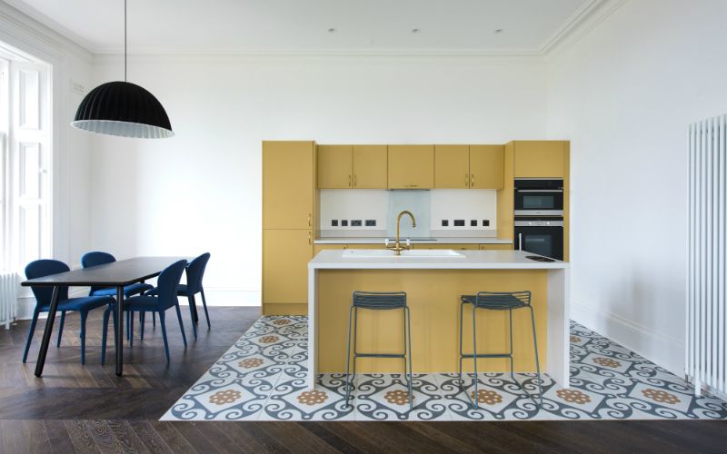 An airy kitchen features a white countertop island paired with two metal stools. Yellow cabinetry provides ample storage, while a sleek dining table with blue chairs occupies the space. A patterned floor adds a distinctive touch, and large windows invite natural light in.