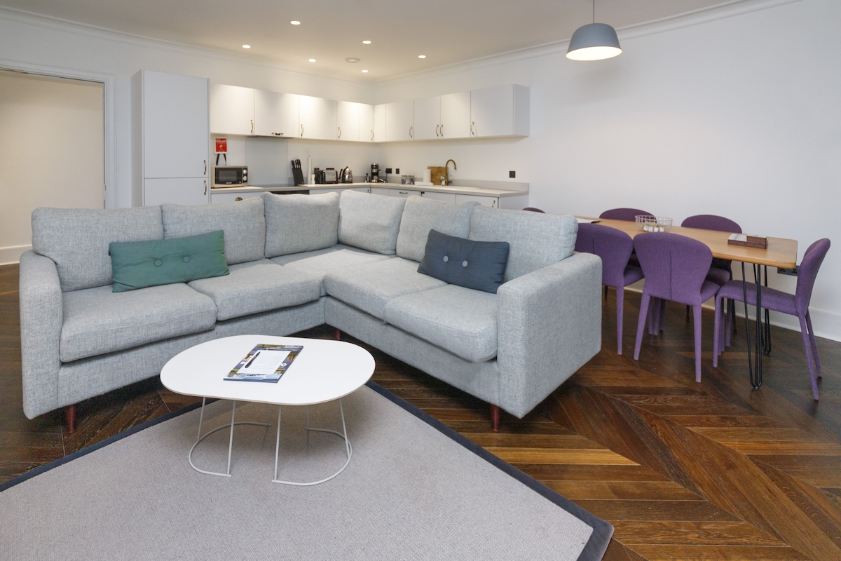 A spacious living area features a light grey sectional sofa, complemented by a circular coffee table. A dining table with purple chairs accommodates six. The fully equipped kitchen is visible in the background, with white cabinetry and modern appliances.