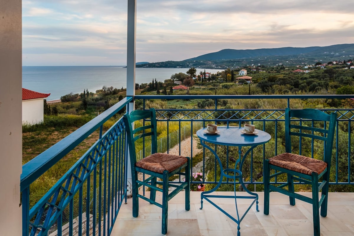 A balcony is depicted with two chairs and a small round table, complemented by coffee cups. The view showcases the sea, rolling hills, and distant greenery under a softly clouded sky, providing a serene atmosphere for relaxation.