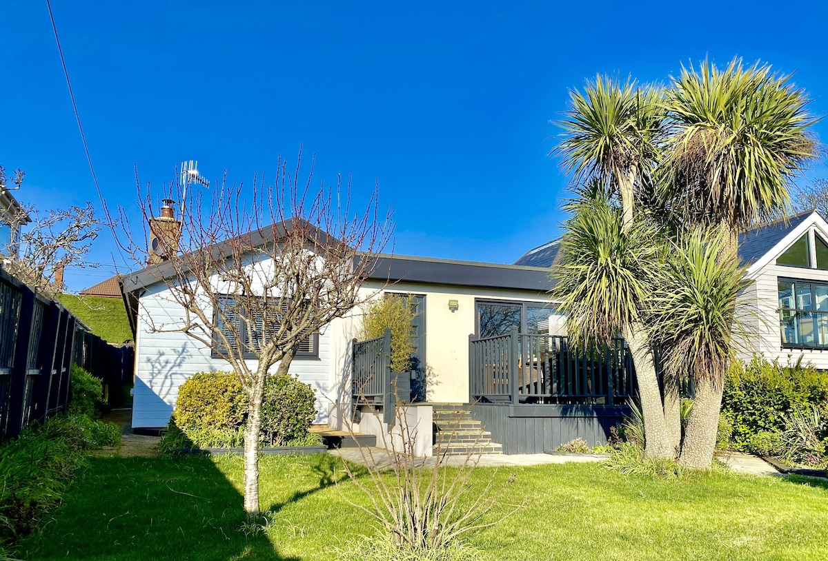 The exterior of the single-storey accommodation is presented with a combination of greenery and well-kept shrubs. A bright blue sky serves as the backdrop, while the entrance features steps leading to a decked area. Nearby, palm trees add a tropical touch to the landscape.