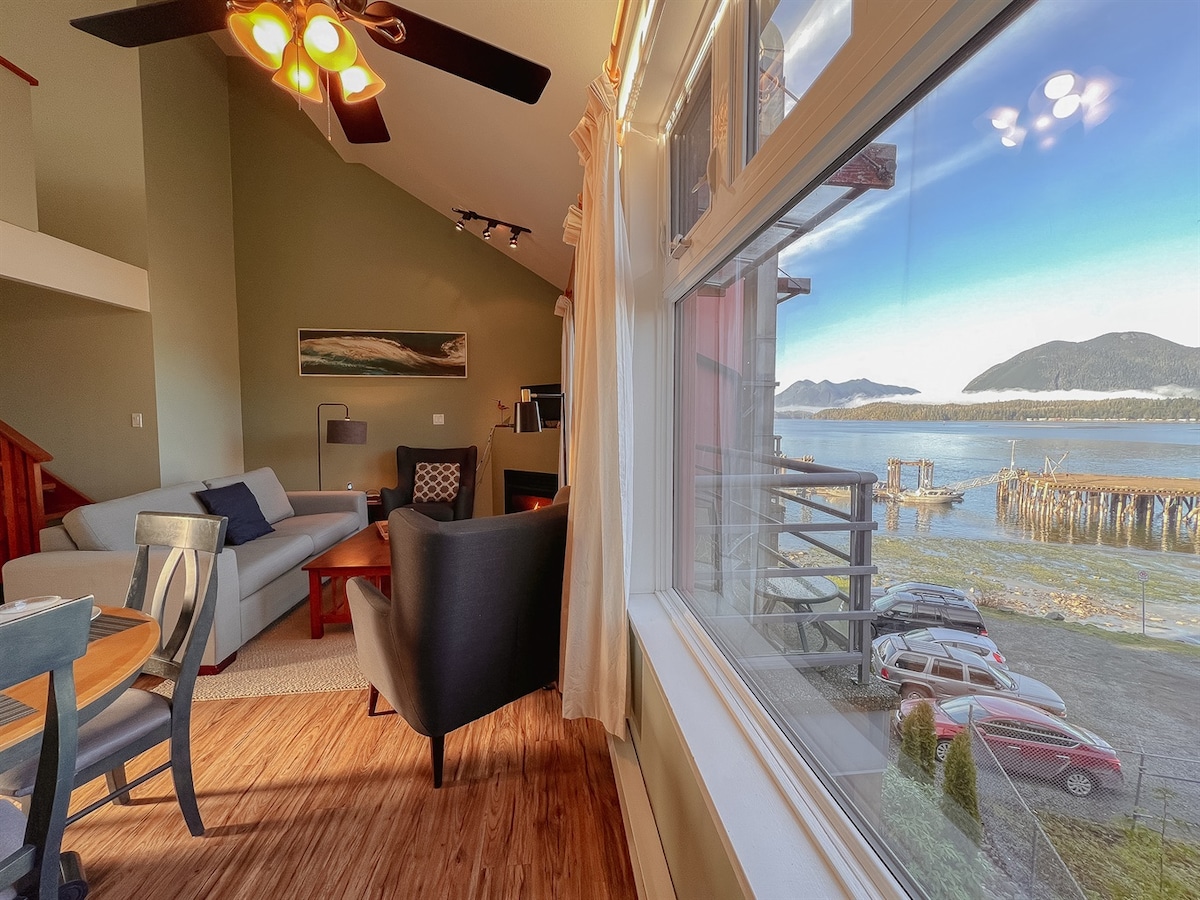 A well-lit open-concept living space is showcased, featuring a cozy couch and a chair. Large windows offer views of the water and distant mountains, while a small dining table accommodates four. The room is complemented by natural light and a warm color palette.