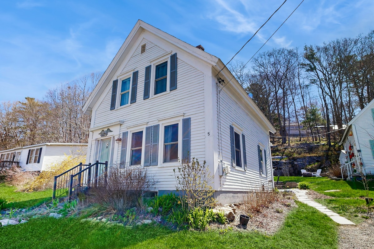 A charming two-story structure features a white exterior with blue shutters. The front yard is landscaped with greenery and colorful plants. Steps lead to the entrance, framed by windows that fill the space with light. The surrounding area showcases wooded hills in the background.