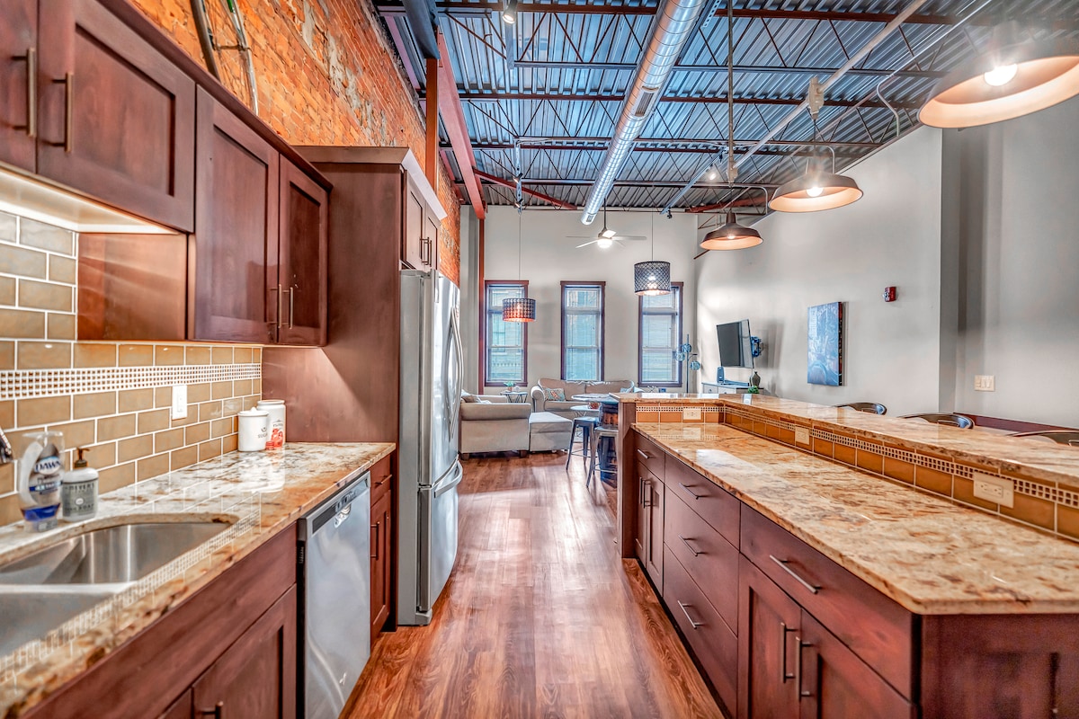 The kitchen features dark wood cabinetry and rich granite countertops, providing ample workspace. Stainless steel appliances are positioned for convenience. The open layout connects to the living area, where natural light filters through large windows, highlighting the industrial-style ceiling with visible beams.