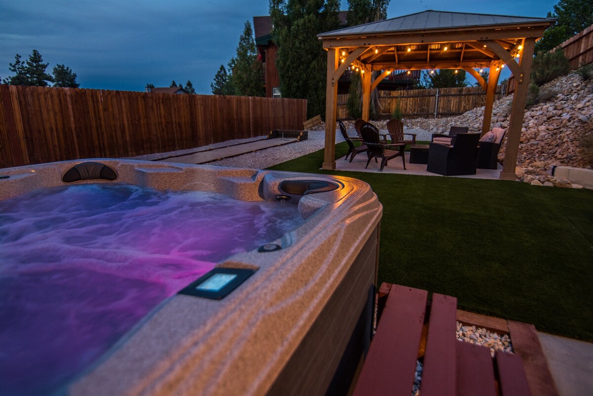 A hot tub is positioned next to a covered gazebo, featuring chairs and a table. Ambient lights are strung along the gazebo's edges, casting a soft glow in the evening. The outdoor space includes a manicured lawn and a gravel pathway bordered by a wooden fence.