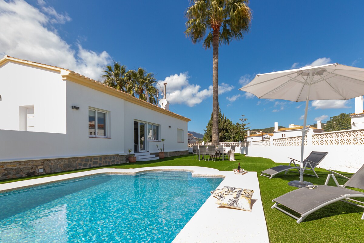 The exterior of the villa features a private pool surrounded by a sunbathing area. Comfortable lounge chairs and a shaded table are positioned on a well-maintained lawn. A tall palm tree adds to the relaxed atmosphere while clear blue skies complement the serene setting.