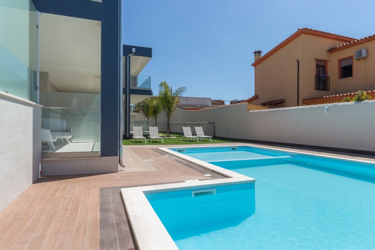 A clear view of the swimming pool is presented, featuring a refreshing blue hue. Sun loungers are arranged along the side, inviting relaxation. The modern apartment building is partially visible in the background, surrounded by greenery and a sunny sky.