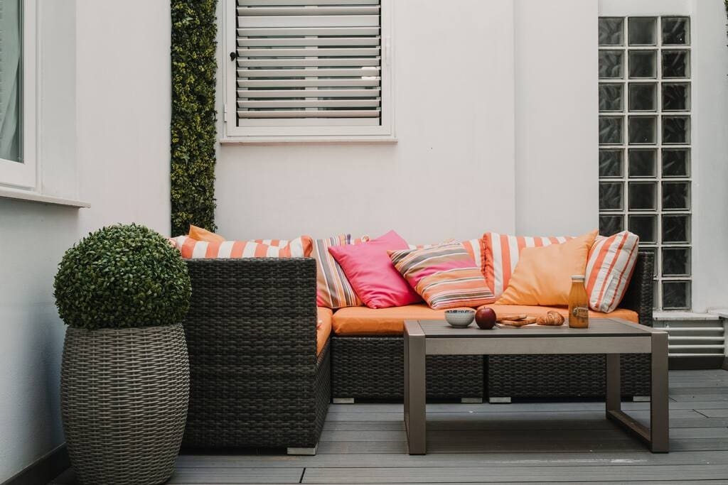 An outdoor seating area features a sectional sofa adorned with inviting orange and striped cushions. A low table sits in front, accompanied by a decorative potted plant. Light-colored walls and a window enhance the bright and open space.
