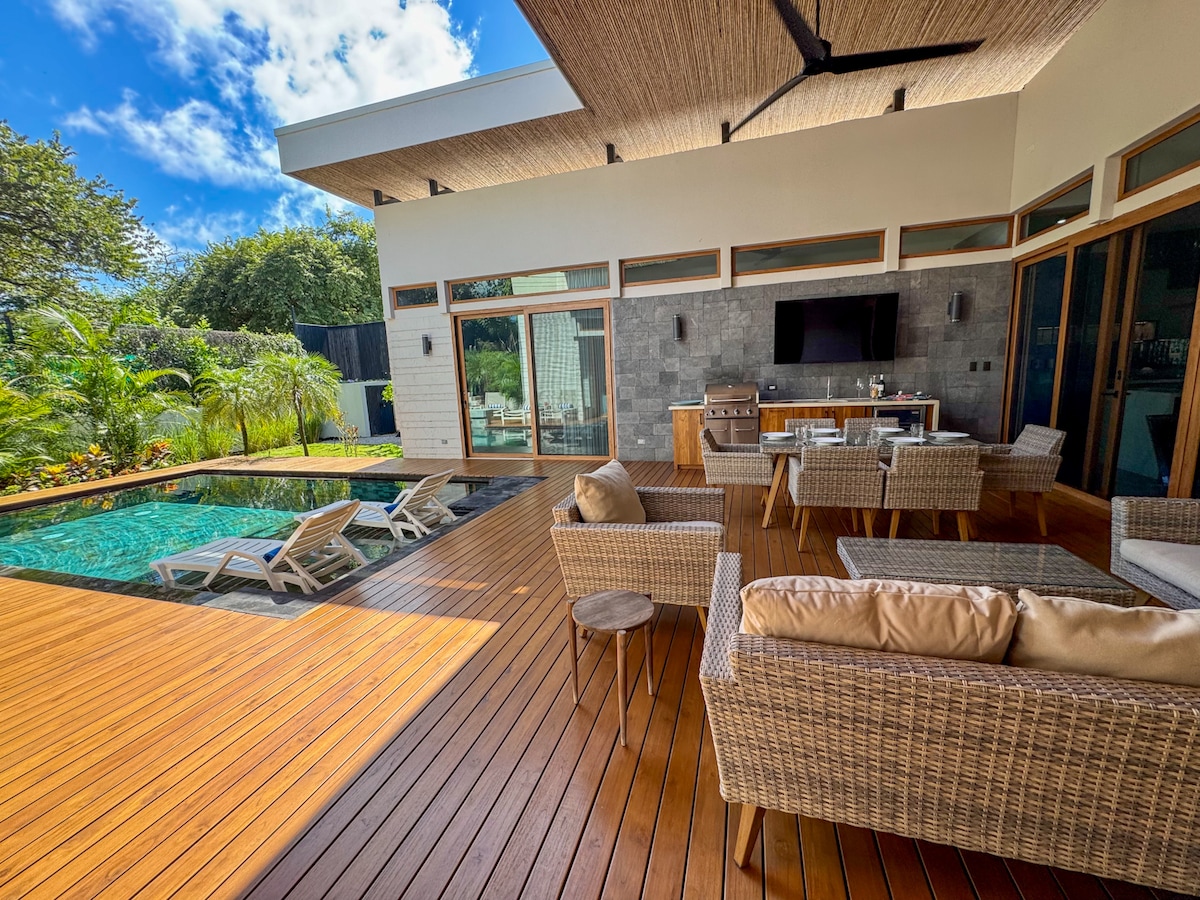 An outdoor living area is seen with comfortable seating surrounding a low table. Lounge chairs are positioned near a private pool, and a fully equipped kitchen is visible in the background. Tall glass doors open to an inviting view of the landscaped area.