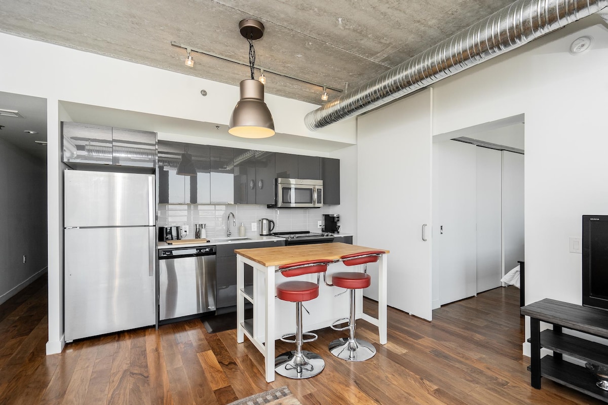 A sleek kitchen area features stainless steel appliances and dark cabinetry. A wooden dining table with red bar stools is placed against the wall, illuminated by overhead lighting. Industrial-style ductwork is visible along the ceiling, complementing the concrete elements.