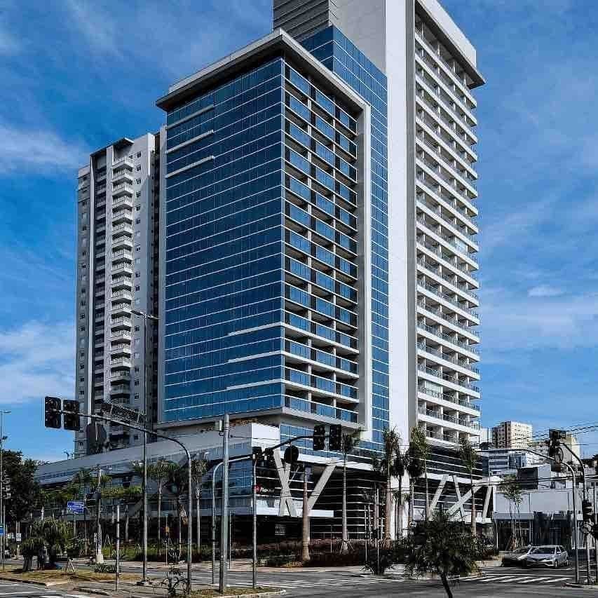 A modern high-rise building features a sleek glass façade, reflecting the sky. The structure is positioned near a bustling intersection, with palm trees lining the street. Pedestrian walkways and traffic signals are visible in the foreground, indicating a vibrant urban setting.