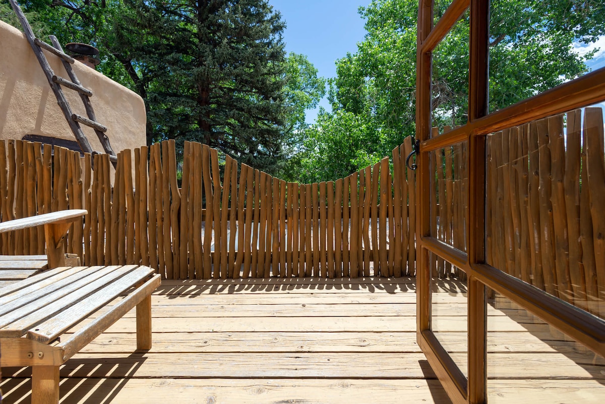 A private balcony features wooden decking and a rustic fence made of natural materials. A wooden chair is positioned for relaxation while providing views of lush greenery and trees surrounding the space. Sunlight streams onto the balcony, creating a bright and inviting atmosphere.