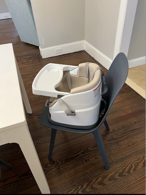 A high chair is seen securely placed on a dark chair, featuring a simple, clean design. The high chair includes a removable tray, and adjustable straps for safety. The surrounding area showcases a wooden floor and neutral-colored walls.