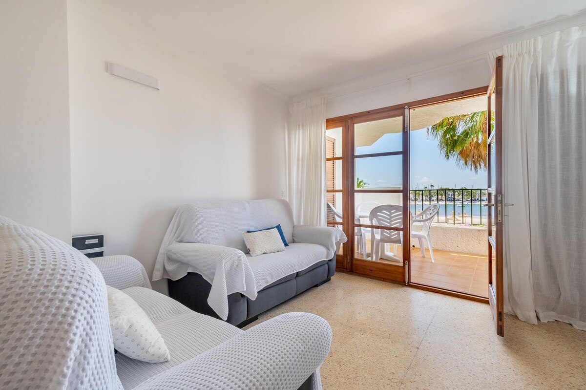A bright living area features a light-colored sofa draped with a soft throw. Large glass doors open to a balcony, allowing natural light and views of the outdoor seating area. White drapes frame the windows, enhancing the airy atmosphere.
