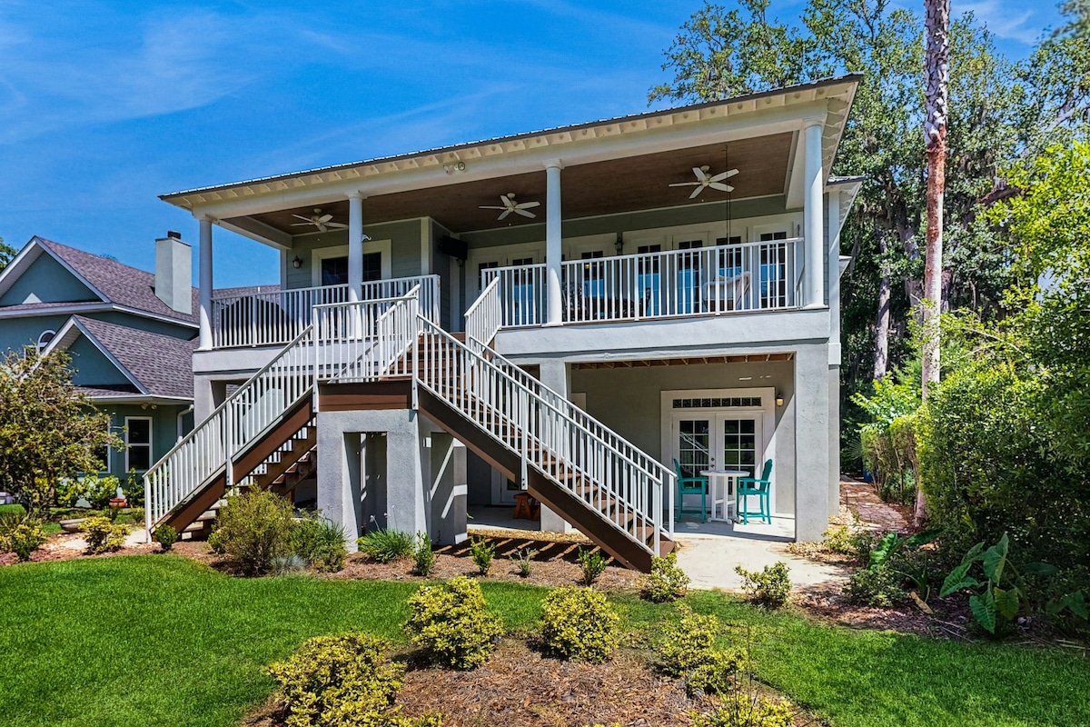 The exterior of a spacious two-story home is shown, featuring a wide staircase leading to a spacious upper deck with ceiling fans. The landscaped yard includes neat shrubbery and greenery, providing a welcoming atmosphere. Natural light enhances the overall appearance of the property.
