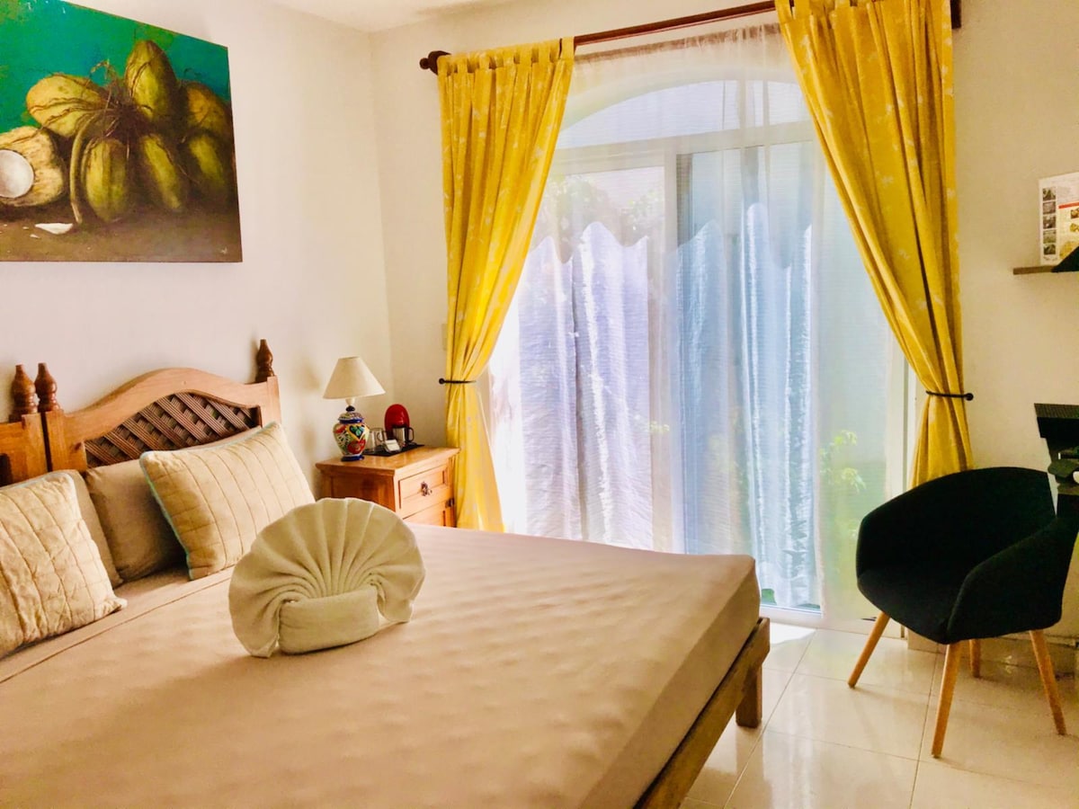 A spacious bedroom is highlighted by a large bed adorned with decorative pillows. Bright yellow curtains frame a doorway leading to the outside, allowing natural light to fill the room. The wooden furniture, including a nightstand and chair, complements the serene atmosphere.