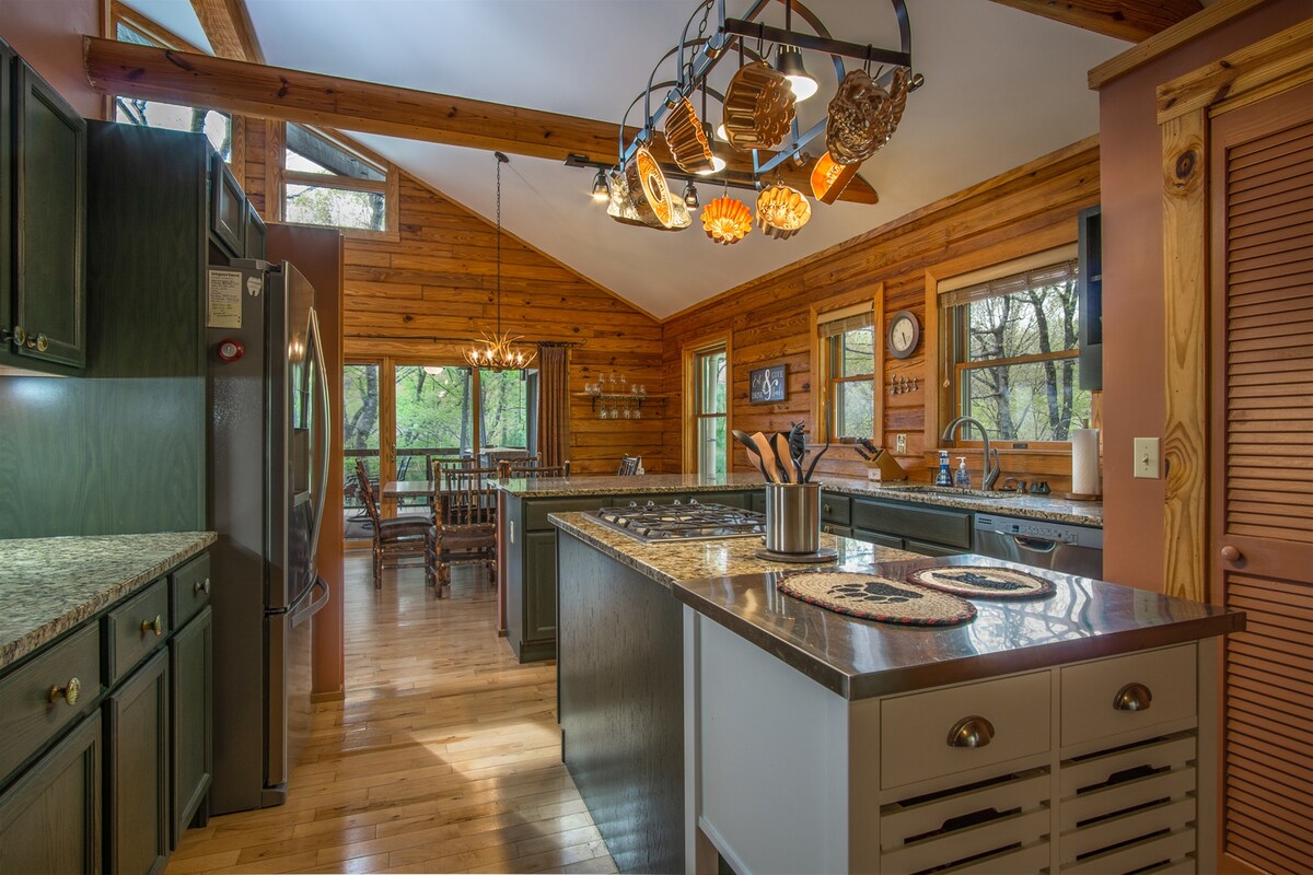 A spacious kitchen features wooden cabinetry and a large island with stainless steel appliances. Natural light floods the space through several windows, highlighting granite countertops and decorative hanging lights. Adjacent is a dining area with a wooden table and chairs, creating a welcoming environment for meals.